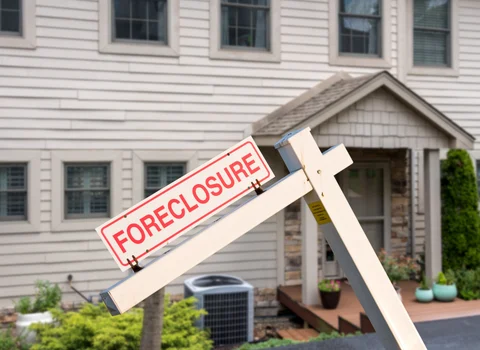 Leaning foreclosure sign in front of tan colonial home with stone entryway in Rhode Island — deed in lieu of foreclosure vs. selling for cash guide from We Pay More Properties